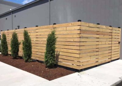 A modern wooden privacy fence encloses an area beside a gray building. Small evergreen shrubs are planted in mulch along the outside of the fence, bordered by a concrete sidewalk.
