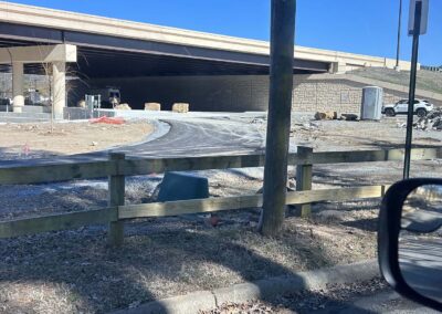 A construction site under a highway overpass with gravel, construction equipment, large rocks, a portable toilet, and a wooden fence in the foreground on a clear day.