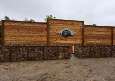 A wooden privacy fence with a sign that reads Patio stands above a stone and metal barrier, with a narrow entrance in the middle and stone paving in the foreground.
