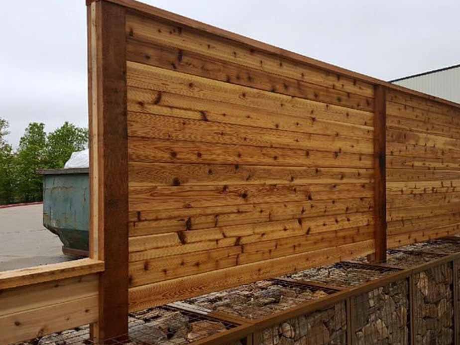A wooden privacy fence is mounted on top of a gabion wall filled with rocks, with trees and a dumpster visible in the background.
