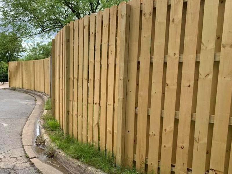 A newly built wooden fence follows the curve of a road, with grass growing alongside the base and trees visible in the background. The street and curb run parallel to the fence.