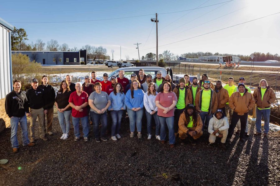 A large group of men and women pose outdoors on gravel, some in work uniforms and safety vests, others in casual clothes, with buildings, vehicles, and trees in the background on a sunny day.