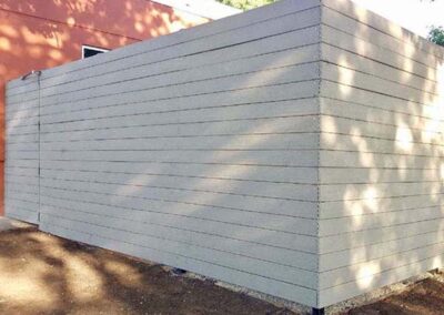 A modern fence made of horizontal concrete panels surrounds a yard, casting soft shadows from nearby trees. The panels are light gray and have a smooth, uniform appearance.