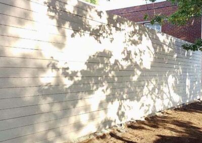Sunlight filters through tree branches, casting dappled leaf shadows onto a long, light-colored wooden fence. The ground beside the fence is bare soil, and part of a brick building is visible in the background.