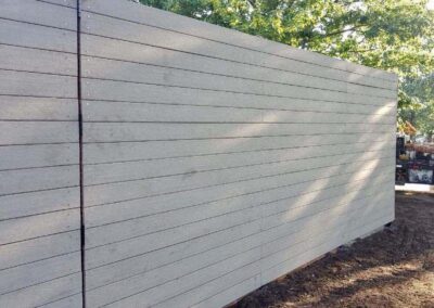 A large wooden fence made of horizontal gray planks stands on bare soil, with green trees and sunlight visible in the background.
