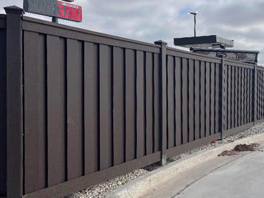 A tall, dark brown privacy fence runs along a sidewalk under a cloudy sky. There is a sign and a light pole visible over the fence in the background. Gravel lies at the base of the fence.