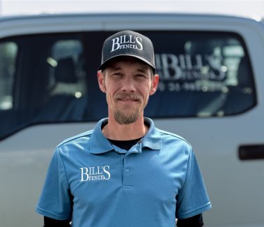 Ed Deemer in a blue polo shirt and black cap with “BILLS FENCE” written on them stands in front of a light-colored truck that also has the “BILLS FENCE” logo on its window.