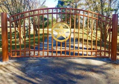 A brown metal gate with vertical bars and a large gold star emblem in the center blocks a tree-lined driveway. Sunlight casts shadows on the ground.