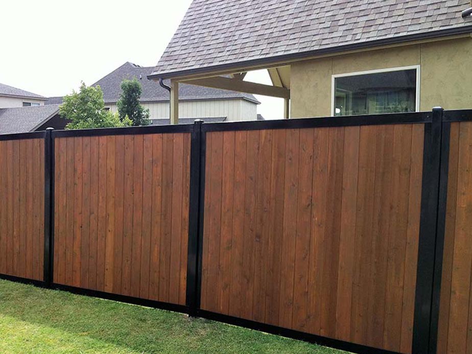A wooden privacy fence with vertical brown panels and black metal posts stands in a grassy yard in front of a beige house with a covered patio and nearby trees.