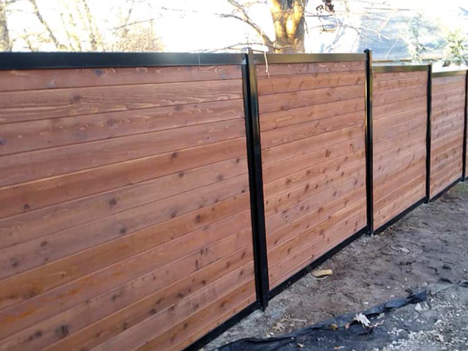A modern wooden privacy fence with horizontal planks and black metal posts runs along a backyard, with sunlight and trees visible in the background.