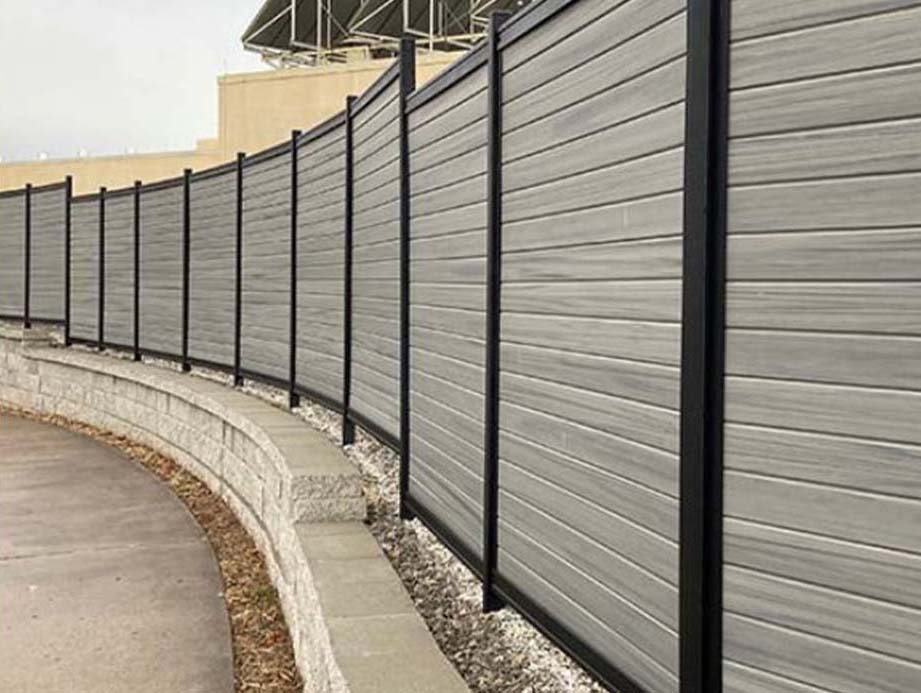 A tall, modern privacy fence with horizontal gray panels and black posts runs alongside a curved concrete retaining wall and a paved walkway. Rocks fill the narrow space between the wall and the fence.
