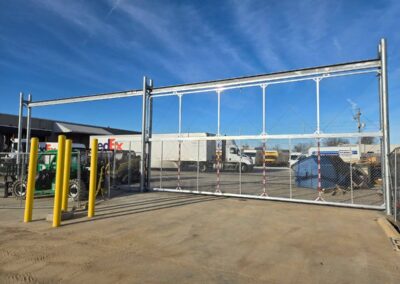A large metal sliding gate with chain-link fencing stands open at an industrial site. Yellow safety bollards are positioned nearby, and delivery trucks, including a FedEx vehicle, are visible in the background.