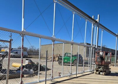 A construction site with a partially built metal framework and chain-link fence. Equipment, tools, and a green forklift are visible nearby, with industrial buildings in the background under a clear blue sky.