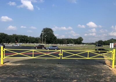 A closed yellow gate blocks a paved road, with several cars driving on a road in the background. Trees and a grassy area stretch out under a blue sky with scattered clouds.