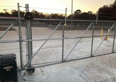 A chain-link sliding gate with barbed wire on top is closed across a paved driveway, with fencing and parking spaces visible in the background at sunset.