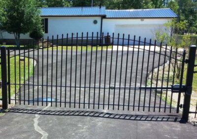 A black metal gate blocks the entrance to a driveway leading to a white house with a blue metal roof and a two-car garage. Trees and grass surround the house, and the driveway is paved with asphalt.