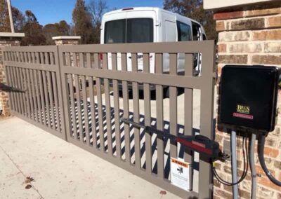 A light gray automatic sliding gate blocks a driveway, with a white van parked behind it. The gate is attached to brick pillars and has a control box on the right side. Trees and a clear sky are in the background.