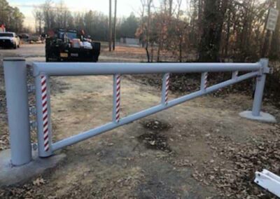 A metal security gate blocks a dirt road in a wooded area. The gate has red and white reflective stripes and is supported by two large cylindrical posts on either end.