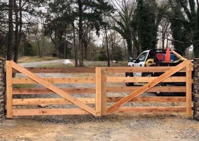 A wooden double gate with diagonal and horizontal planks stands between two stone pillars. Behind the gate, a white pickup truck is parked on a gravel driveway with trees in the background.