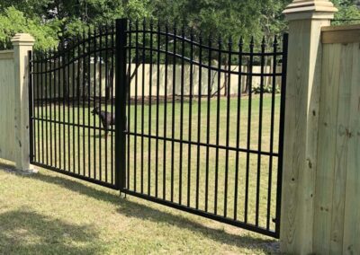A black metal gate with vertical bars and pointed tops is attached to wooden posts, enclosing a grassy yard with a dog visible in the background. Trees and a wooden fence are in the distance.