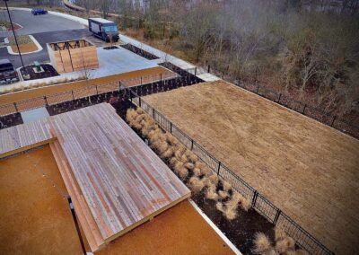 Aerial view of a landscaped outdoor area with a wooden deck, pathways, plant beds, and a grassy section bordered by a black fence. A parking lot with a truck and a wooded area are in the background.