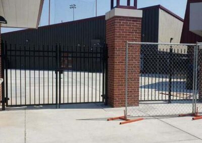 A chain-link fence block stands next to an open gate, creating an unnecessary barrier in front of an unobstructed entrance to a building with metal siding and brick columns.