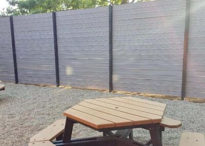 A hexagonal wooden picnic table with benches sits on gravel in front of a tall, modern privacy fence on a sunny day. Trees are visible above the fence in the background.