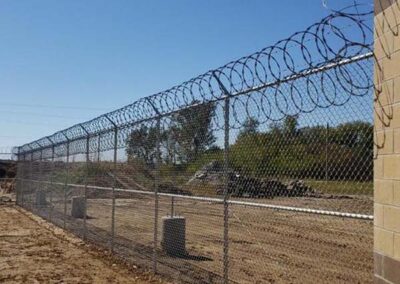A tall chain-link fence topped with barbed wire and razor wire surrounds a dirt area, with trees and vegetation in the background under a clear blue sky.