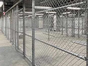 A large, empty indoor space divided by metal chain-link fences, forming multiple cage-like enclosures under fluorescent lights. The area appears industrial and unoccupied.