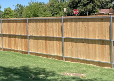 A tall wooden privacy fence with metal supports stands on a green lawn, with a stop sign visible in the background and trees surrounding the area.