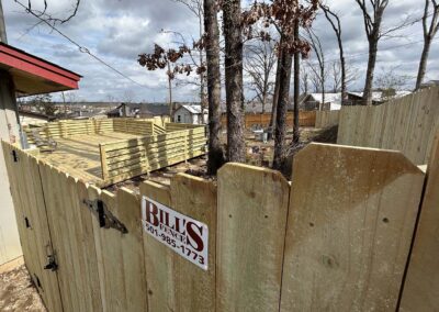 A newly built wooden fence surrounds a deck area in a backyard. Trees without leaves stand inside the fenced area. A sign on the fence reads BILLS FENCE along with a phone number. Houses are visible in the background.