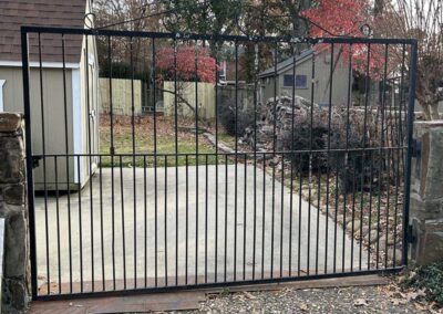 A black iron gate blocks the entrance to a driveway leading to a shed and backyard. Trees with autumn leaves and scattered branches are visible in the fenced yard.