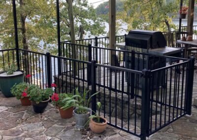 A black metal fence encloses a grill on a stone patio overlooking a lake, with potted plants and flowers arranged outside the fence and trees in the background.