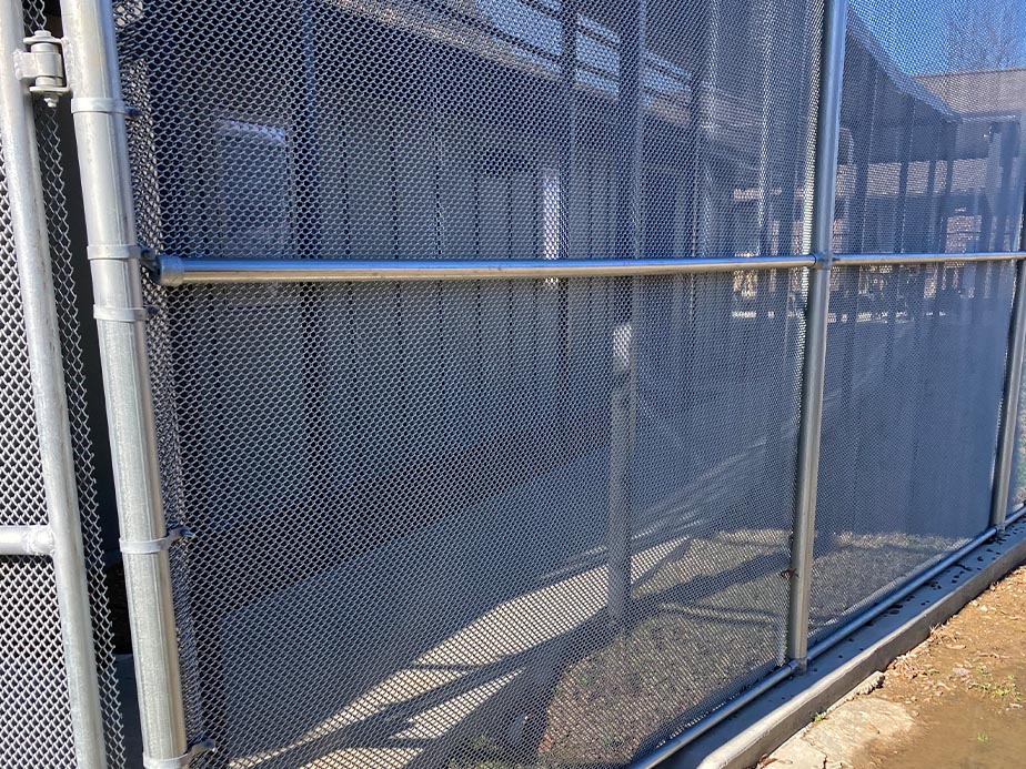 A tall metal mesh fence with metal poles and a shadow on the ground, partially obscuring a building and walkway behind it on a sunny day.