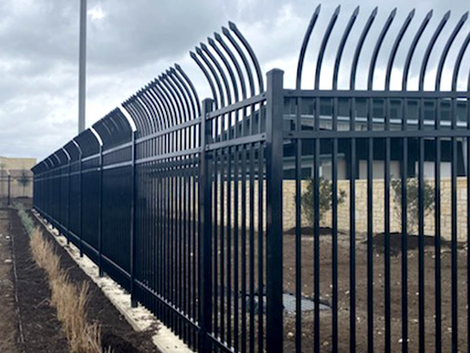 A tall black metal security fence with curved spikes on top lines a dirt area under a cloudy sky, providing a barrier alongside a building and road.