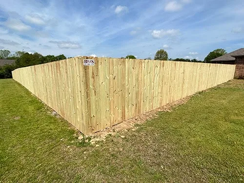A tall wooden privacy fence encloses a grassy yard, forming a sharp corner. The fence is made of vertical planks, and a small sign is attached near the corner. A house and trees are visible in the background.
