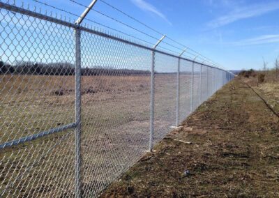 A long chain-link fence topped with barbed wire stretches across a field, separating grassy land on one side from open terrain on the other under a clear blue sky.