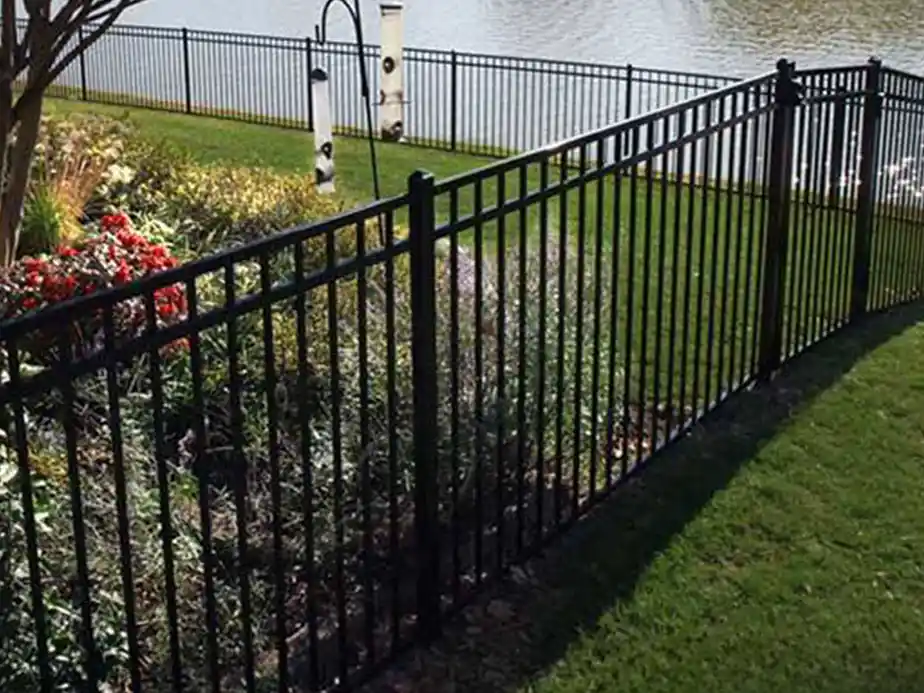 A black metal fence encloses a garden bed with green grass, colorful flowers, and bushes. Beyond the fence is more grass, bird feeders, and a pond with another matching fence along the water’s edge.