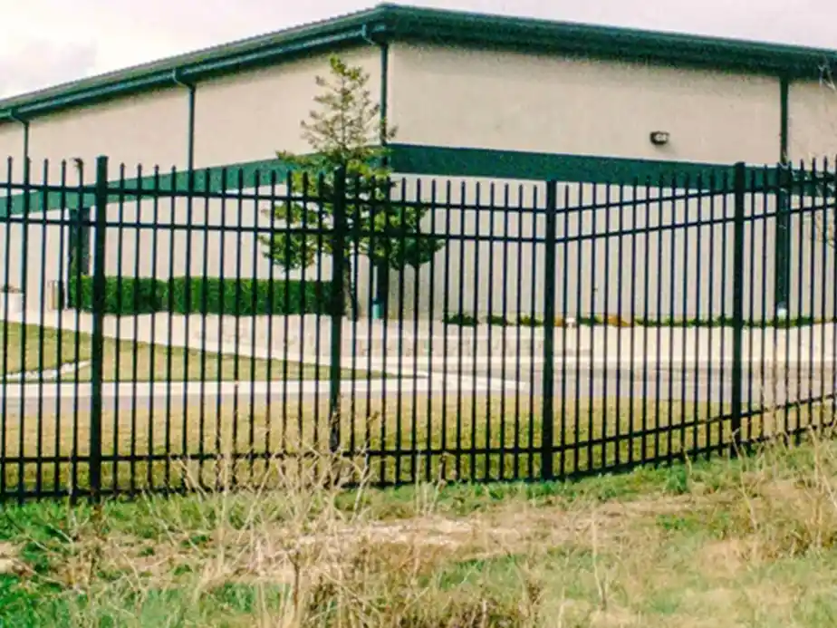 A black metal fence is bent and warped in front of a beige industrial building with green trim. There is a small tree and a sidewalk inside the fenced area, with grass and weeds in the foreground.