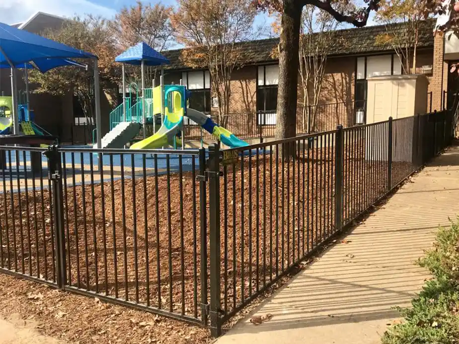 A fenced playground with a blue and green slide and climbing structure sits beside a building, surrounded by trees with autumn leaves and a pathway leading to the gate.