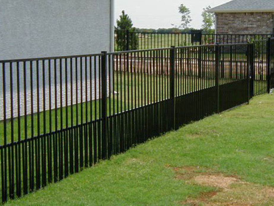 Black metal fence with vertical bars divides a grassy yard, running alongside houses. The fence appears sturdy and well-maintained, with green lawn on both sides and a few patches of bare earth.