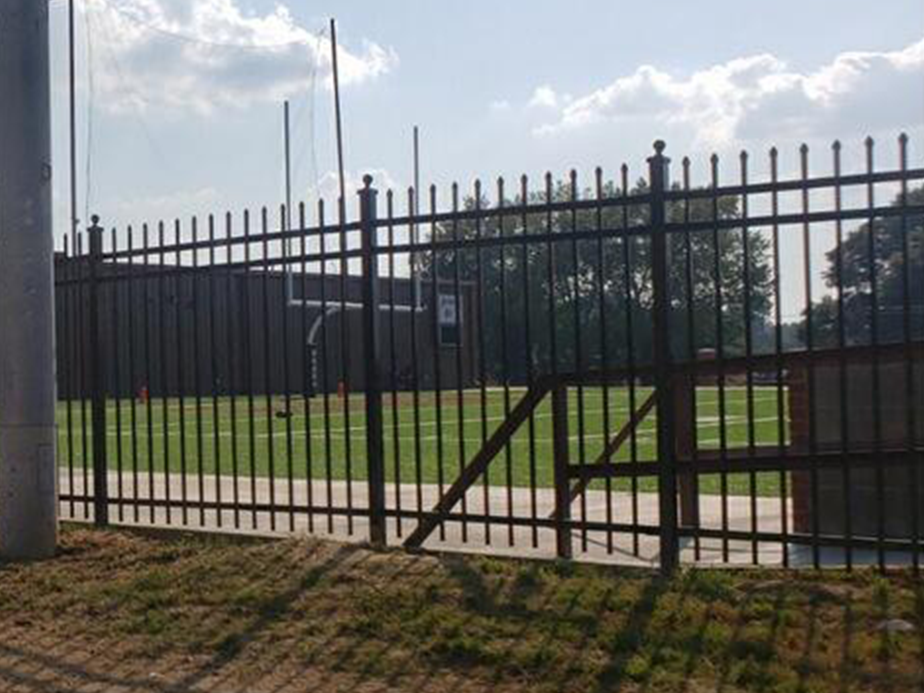 A metal fence with a broken gate surrounds a green outdoor sports field. Tall goalposts rise in the background beneath a partly cloudy sky.