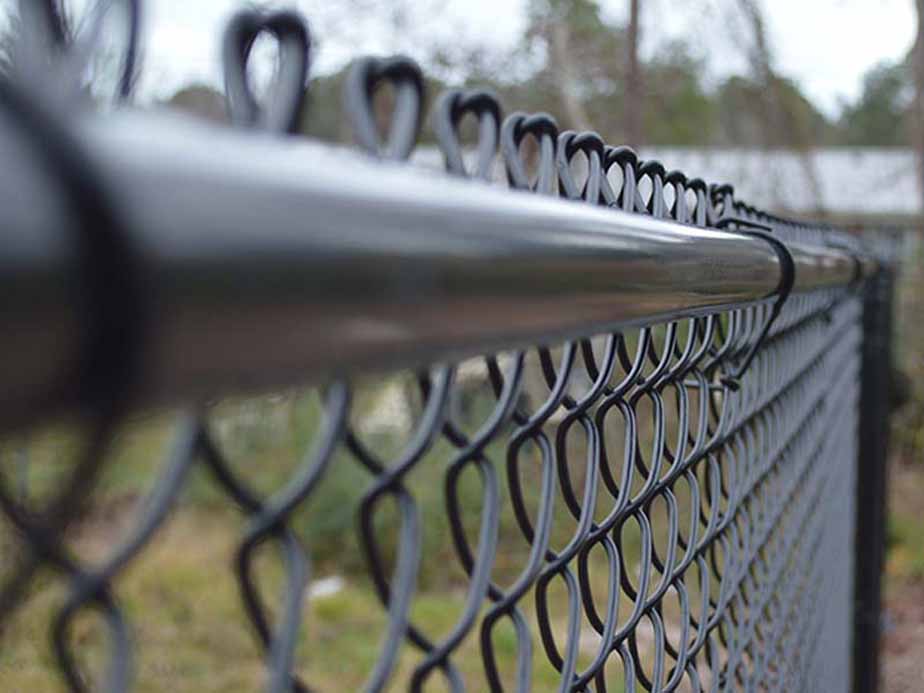Close-up view of a black chain link fence, with the focus on the top rail and the repeating wire pattern. The background is blurred but shows greenery and trees.