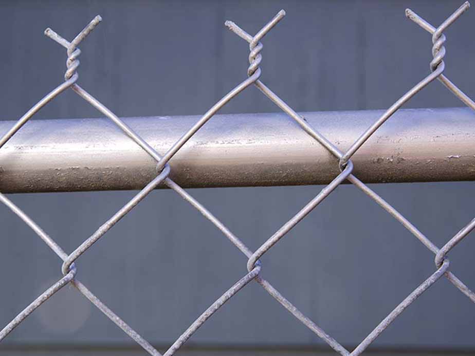 Close-up view of a metal chain-link fence with a horizontal metal pipe running behind it, against a plain, blurred gray background.