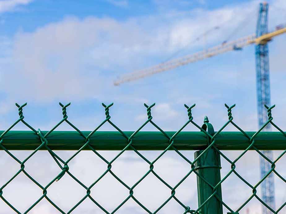 Green metal chain-link fence with barbed wire on top, set against a blue sky with clouds. A tall construction crane is visible in the blurred background.