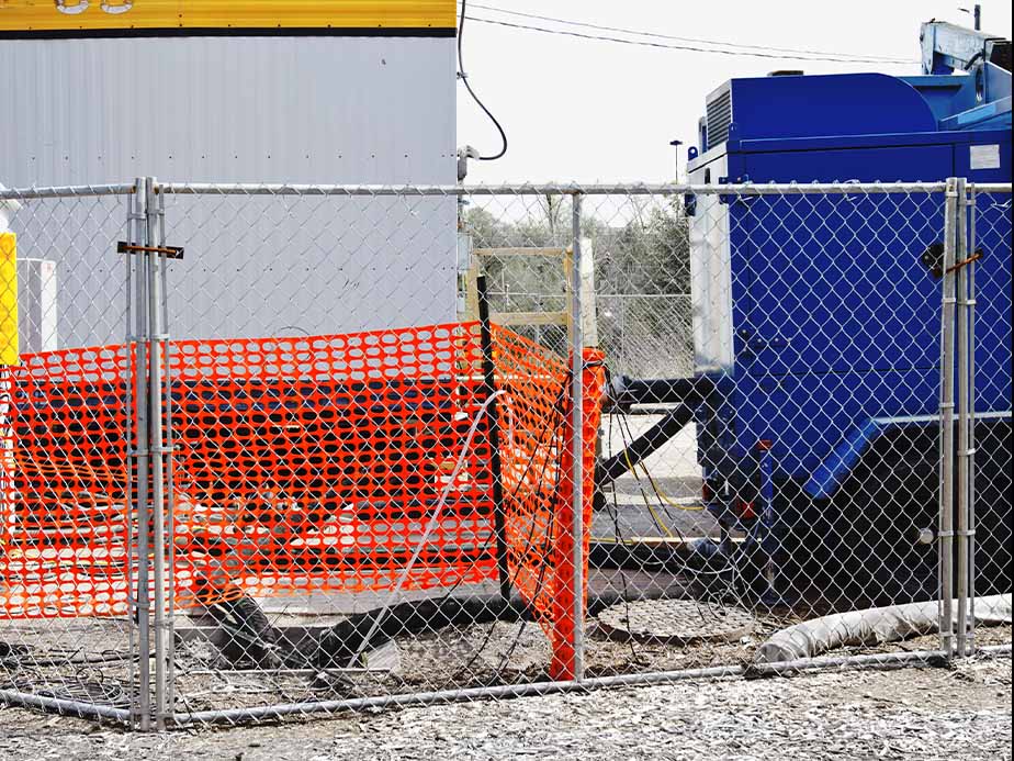 A construction site with a chain-link fence and orange safety netting surrounds equipment and debris. A blue industrial machine and a white trailer are visible in the background.