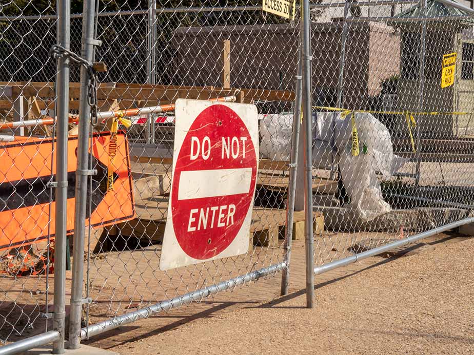 A Do Not Enter sign is attached to a chain-link fence blocking access to a construction site with various materials and equipment visible behind the fence.