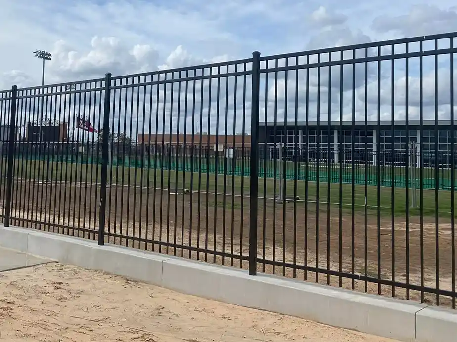 A black metal fence encloses a grassy athletic field with a sand path in the foreground and buildings visible in the background under a partly cloudy sky.