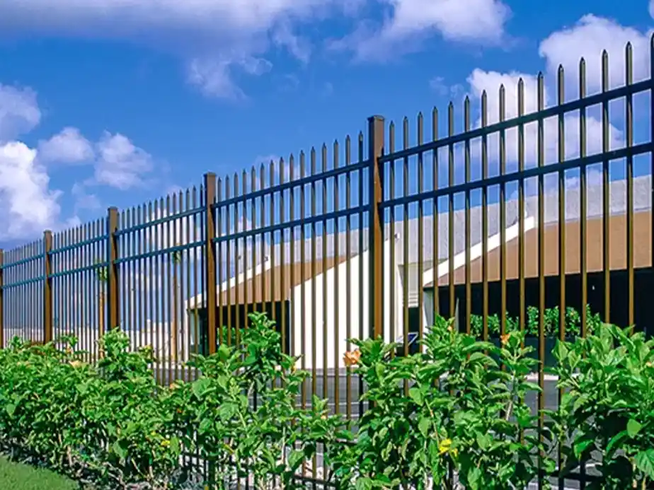 A tall metal fence with vertical bars and pointed tips runs along a property, with green shrubs growing in front. A modern building with angled roofs is visible behind the fence under a blue sky with scattered clouds.