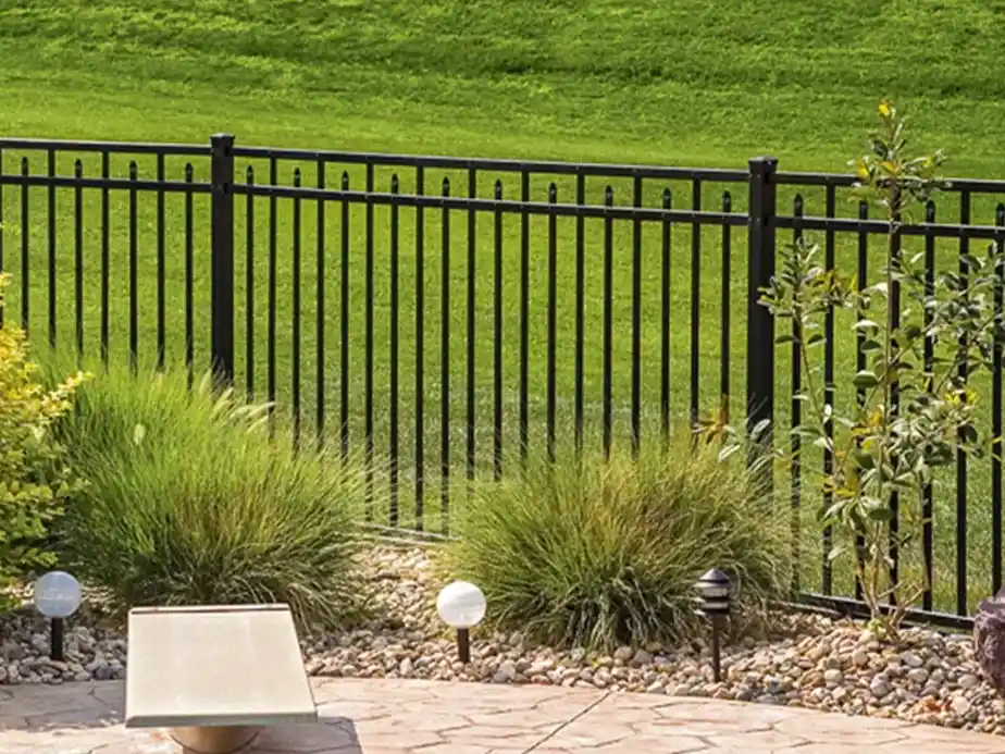 Black metal fence with vertical bars borders a neatly trimmed green lawn. In front, there are decorative grasses, small plants, pathway lights, and a stone patio area.